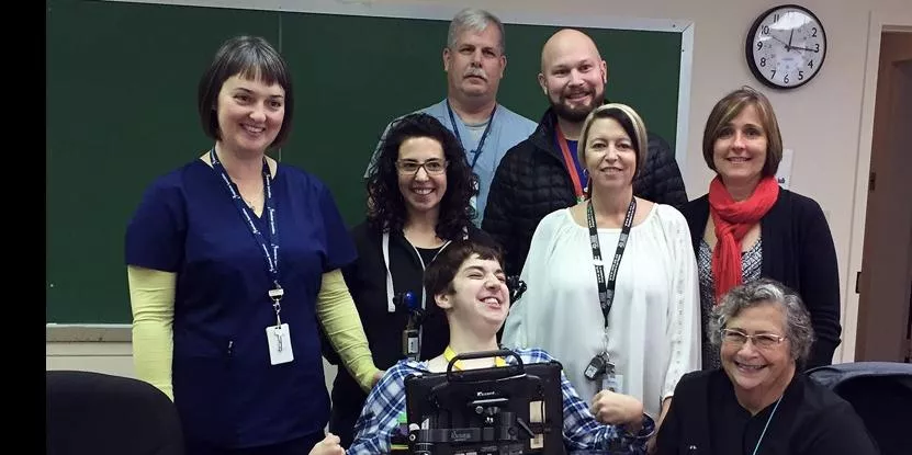 Group of eight people in a room with a chalkboard and clock, one using a communication device.
