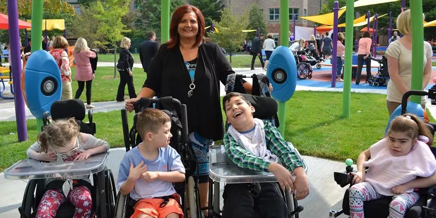 Woman stands behind four children in wheelchairs at an outdoor playground with colorful equipment.