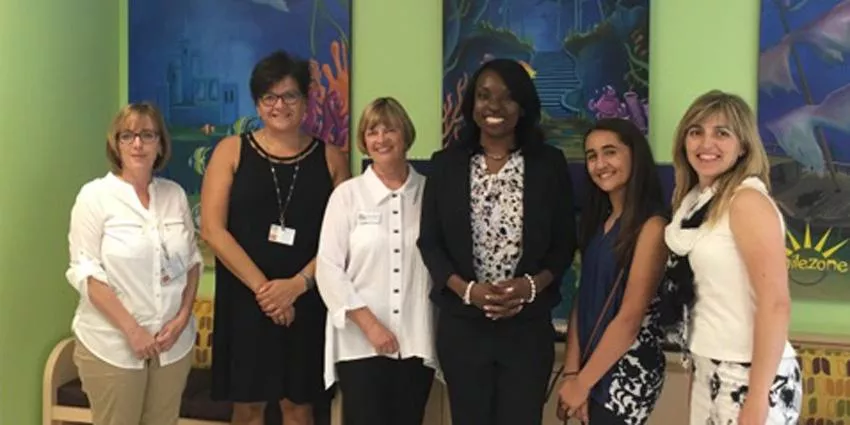 Six women in business casual attire standing indoors against a colorful painted wall.