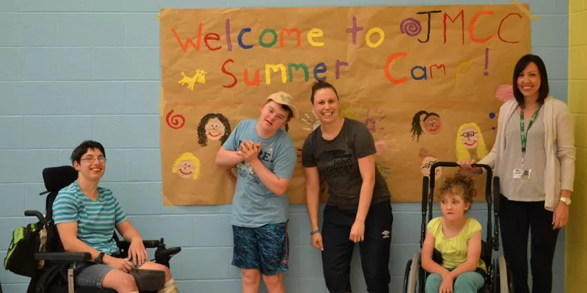 Five people, including two in wheelchairs, stand in front of a JMCC Summer Camp sign.