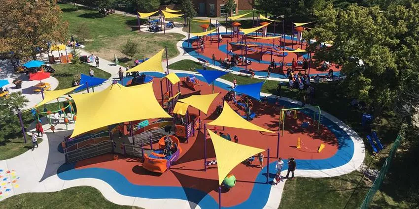 Playground with yellow and blue shade sails, red and blue surfaces, pathways, and people.