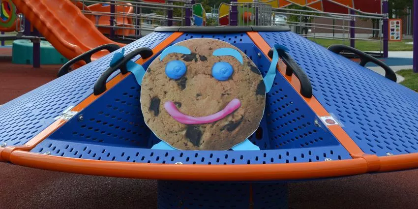 Playground climbing structure with cookie face design, blue and orange colors, and nearby slide.