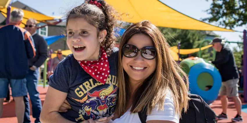 Woman wearing sunglasses holding smiling young girl with curly hair and red bandana at playground.