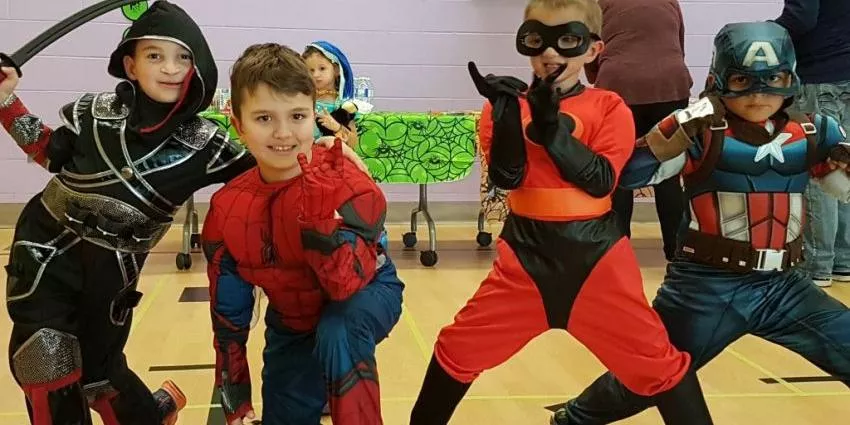 Four children in superhero and action character costumes posing on a gymnasium floor.