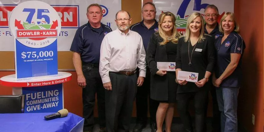 Seven people standing by a 75th anniversary Dowler-Karn sign and holding envelopes indoors.