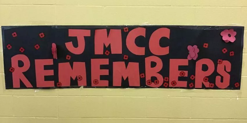 Banner with red letters JMCC REMEMBERS on black background decorated with small red and pink flowers.