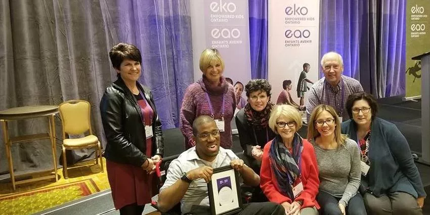 Group of eight people posing indoors with eko Empowered Kids Ontario banners in background.