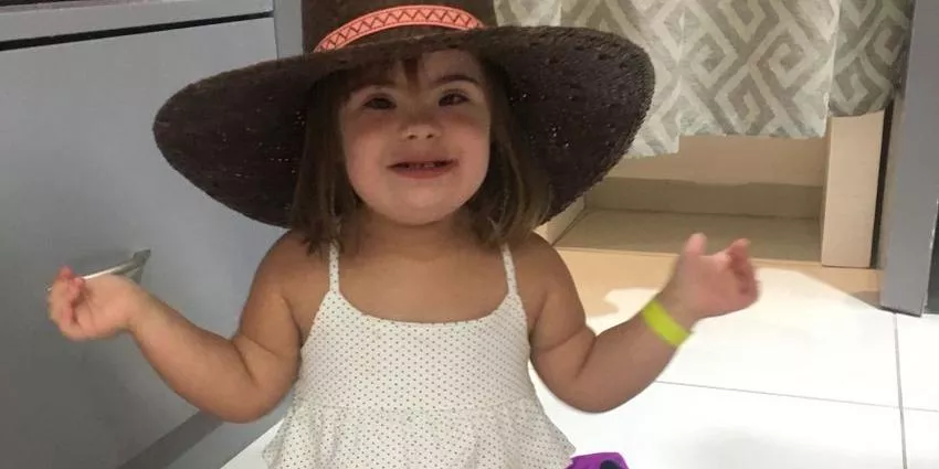 Toddler in a white polka dot dress and large woven hat standing indoors near a patterned curtain.