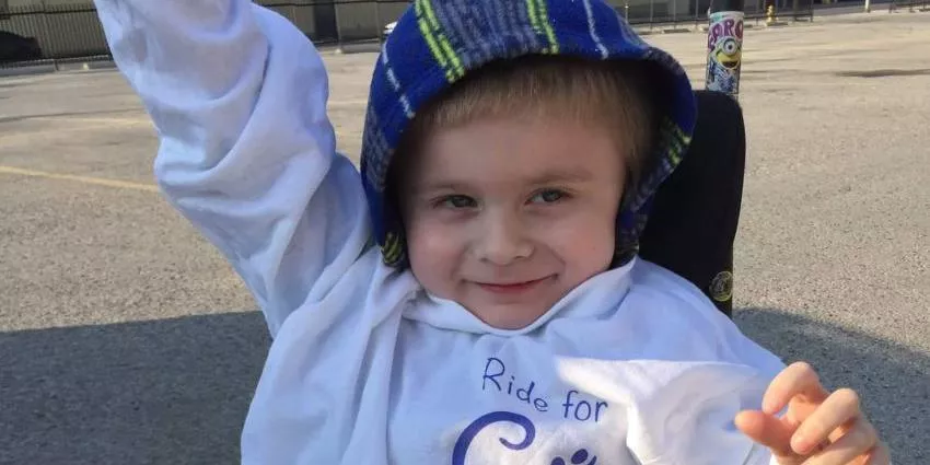 Boy wearing a blue plaid hat and white hoodie outdoors raising one arm and smiling.