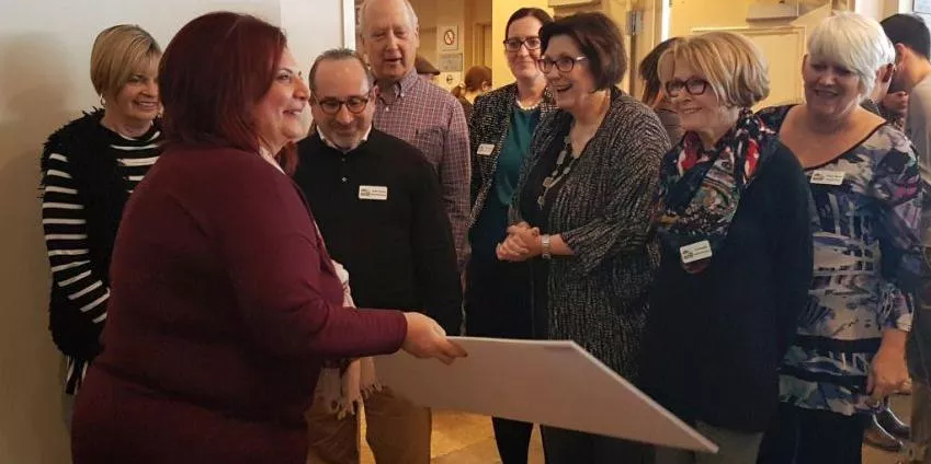Group of eight people indoors, one woman holding a large board, others smiling and wearing name tags.