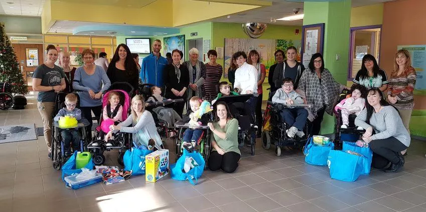 Group of adults and children, some in wheelchairs, with blue bags and toys in a colorful room.