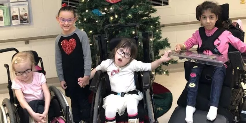 Four children, two standing and two in wheelchairs, holding hands in front of a decorated Christmas tree.
