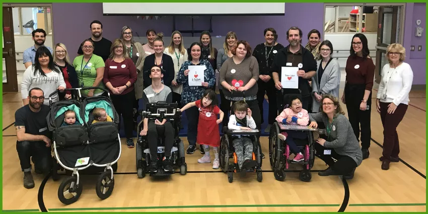 Group photo of adults and children, some in wheelchairs and strollers, in a gymnasium event.