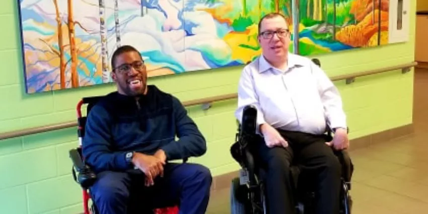 Two men in wheelchairs sit indoors in front of colorful nature-themed wall art.