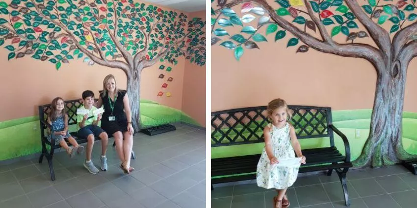 Room with mural of a large tree, black metal bench, tiled floor, and children with an adult.