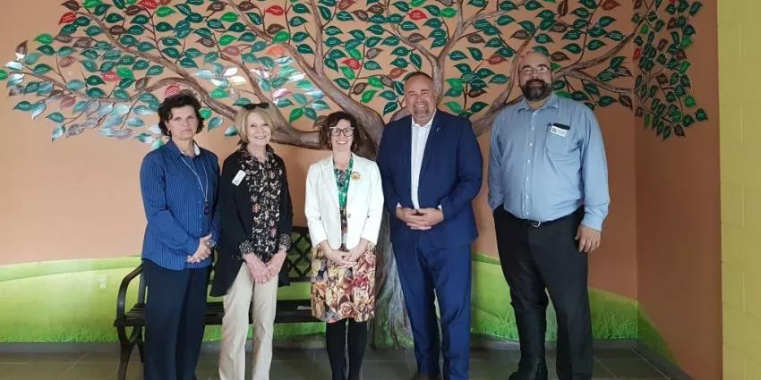 Five people standing indoors in front of a wall mural of a tree with green leaves.