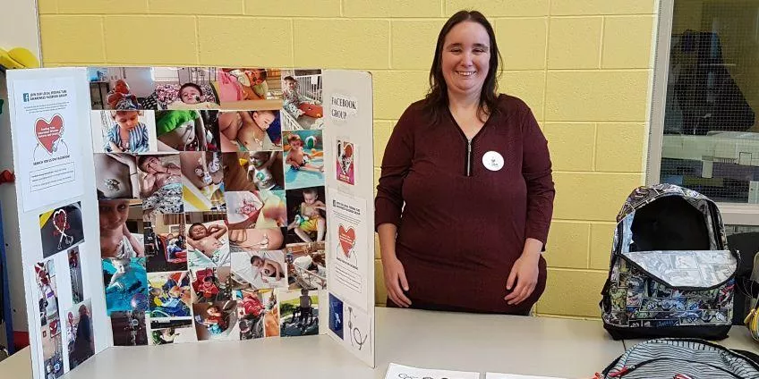 Woman stands behind table with photo collage display board and two bags in indoor setting.