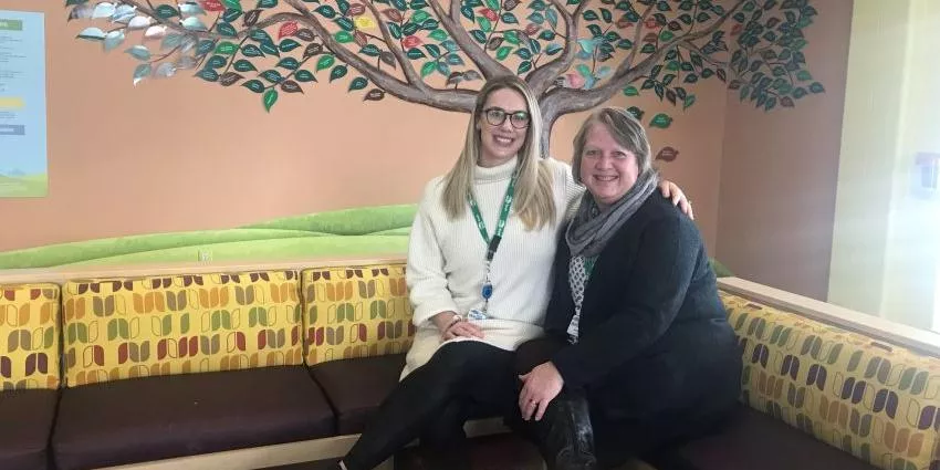 Two women seated on a patterned bench in front of a colorful tree mural on the wall.