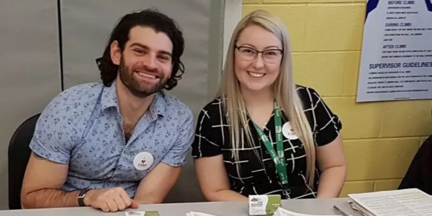 Two people sitting at a table with papers and informational materials against a yellow wall.