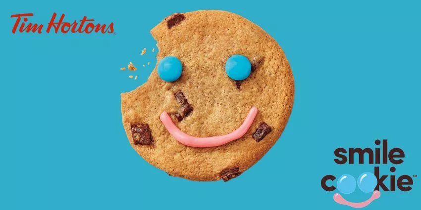 Partially eaten chocolate chip smile cookie with blue candies eyes and pink icing smile on blue background.