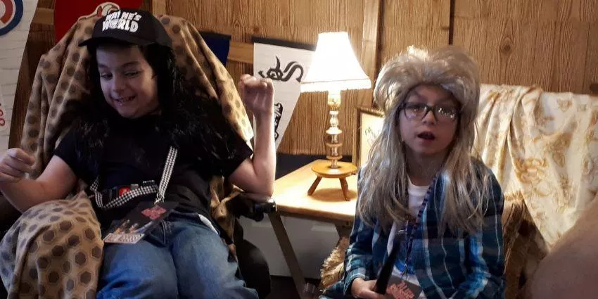 Two children in wigs and casual clothes sit on patterned chairs in a wood-paneled room.