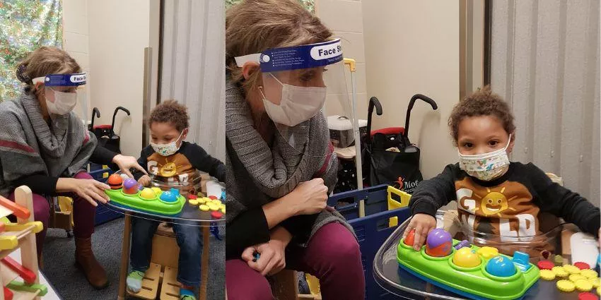Woman with face shield and mask sitting next to masked child playing with toy on clear table indoors