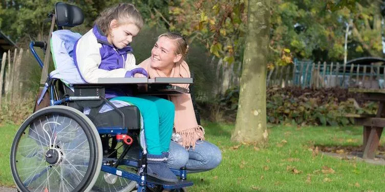 Young woman in wheelchair outdoors with another woman kneeling beside her on grass.
