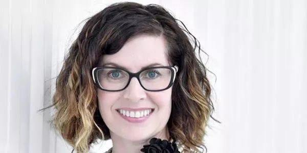Woman with shoulder-length wavy hair, black glasses, and light top smiling against white background