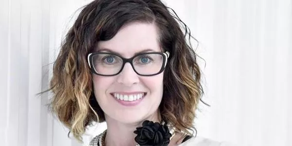 Woman with short curly brown hair and black-rimmed glasses wearing a white top with black flower accessory.
