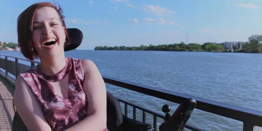 Person in wheelchair laughing by railing near large body of water under a partly cloudy sky