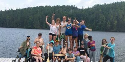 Group of people posing on a dock by a lake with forest in the background, some standing and some seated.