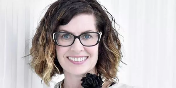 Woman with wavy shoulder-length hair, black-framed glasses, and a white top with a black flower accessory smiling against a light background