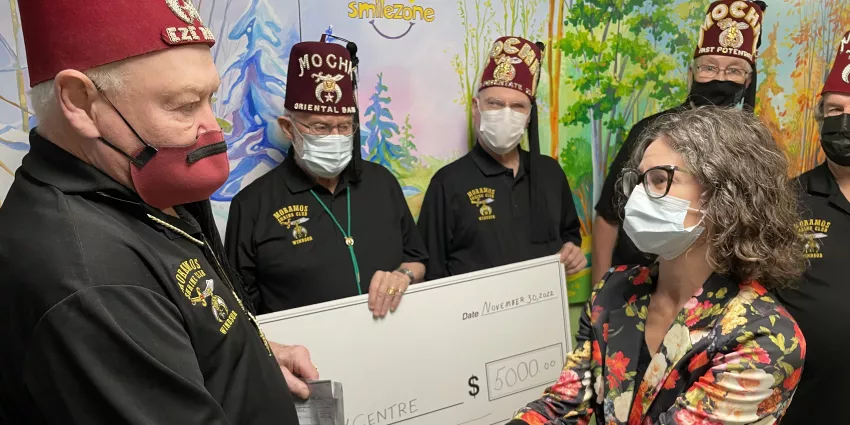 Group wearing matching shirts and red fezzes presents $5000 check to masked woman indoors.
