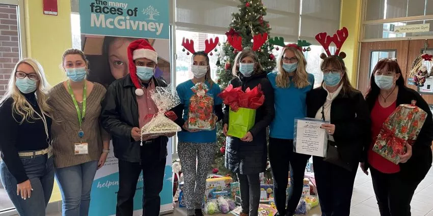 Eight masked people in festive attire hold gifts near a Christmas tree indoors.