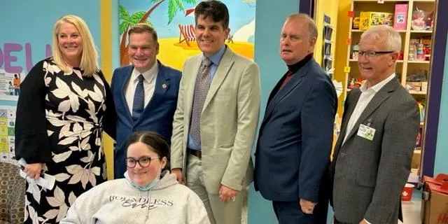 Five adults standing behind a seated young person with glasses in a colorful room with a beach mural.
