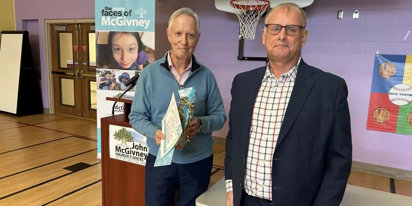 Two men stand in a gymnasium near a podium with John McGivney Children's Centre signage.