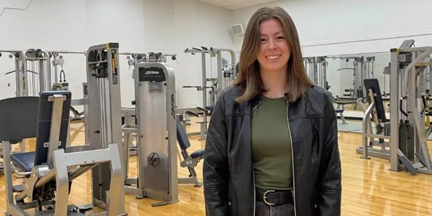 Woman standing in gym with exercise machines, mirrors, and wooden floor background.