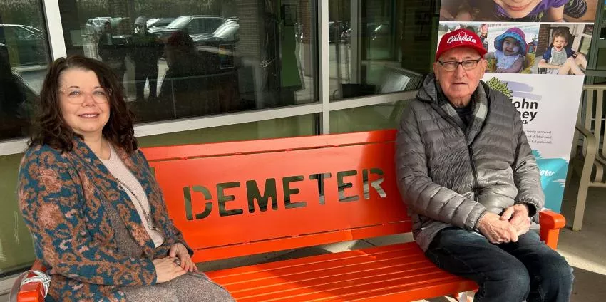 A woman and elderly man seated on an orange bench with DEMETER cut out on backrest.
