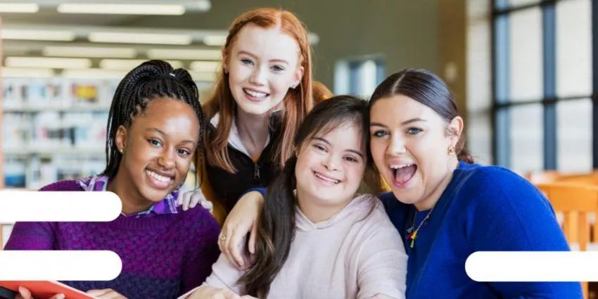 Four young women sitting together in a library, smiling with bookshelves and windows in background