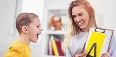Woman holding clipboard with letter A smiling at boy in a bright room with shelves.