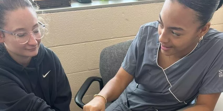 Medical professional in scrubs assisting a person wearing glasses and a dark hoodie.