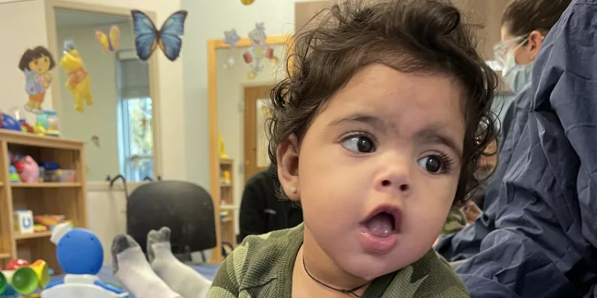 Young child with curly hair in a green outfit seated in a playroom with adults wearing masks.