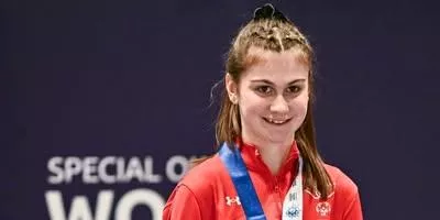 Female athlete wearing red jacket with blue medal ribbon standing by event backdrop.