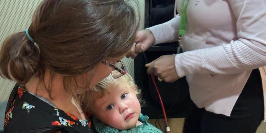 Woman with glasses holds a toddler wrapped in a green blanket while another person manages cables nearby