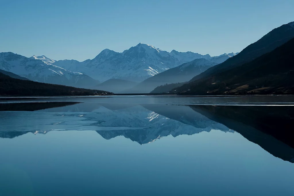 Lake with clear reflections of snow-covered mountains and forested hills under a blue sky.