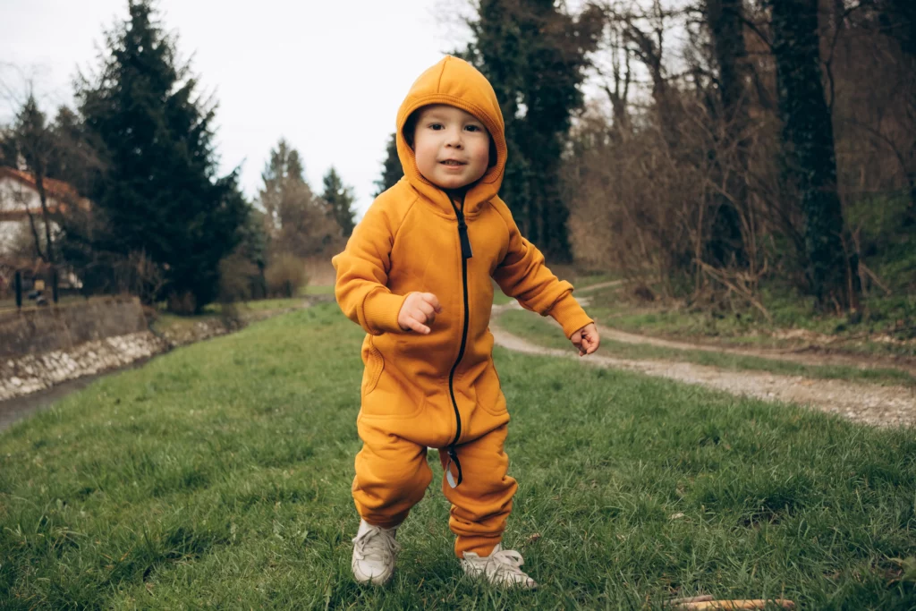 Toddler in orange hooded jumpsuit and white sneakers walking on grassy outdoor path near trees.