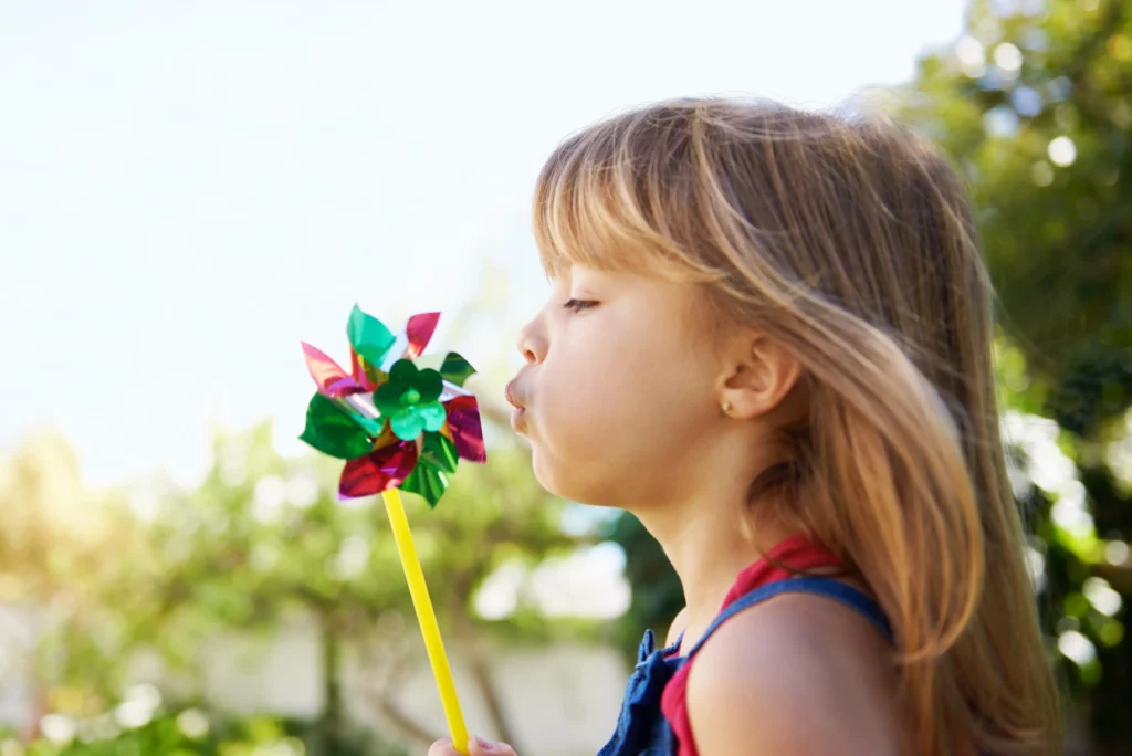 Young girl outdoors blowing on a colorful pinwheel on a sunny day.