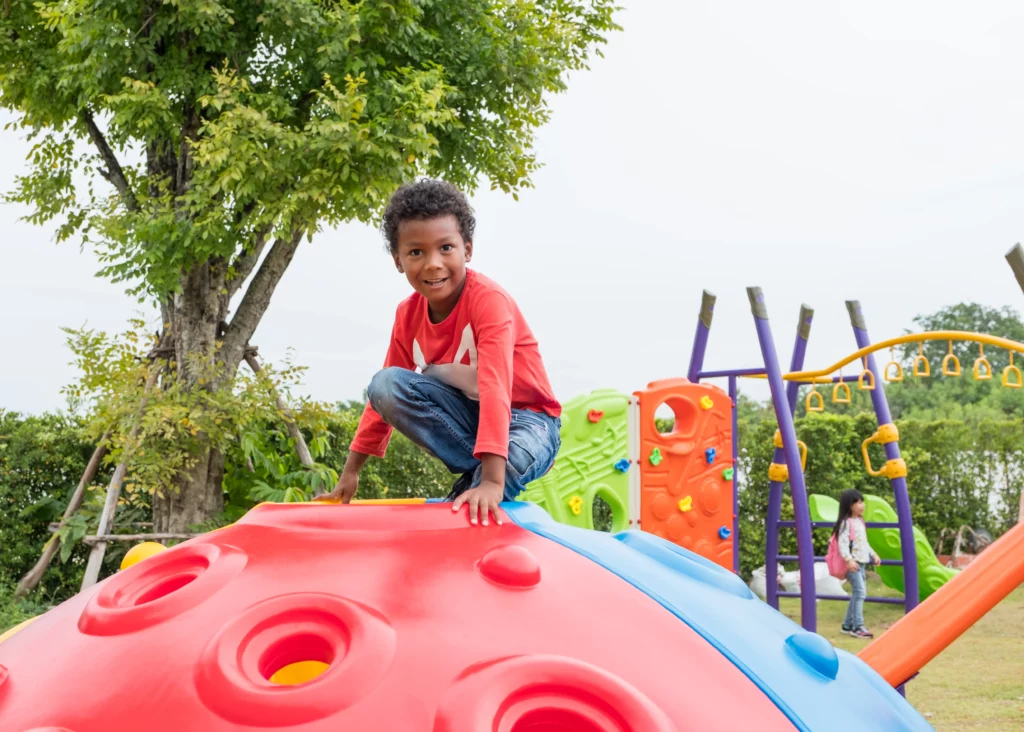 Child in red shirt climbing a colorful playground structure with other play equipment and children nearby.