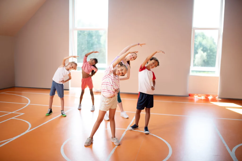 Children stretching in a gymnasium with an orange floor during a group exercise session.