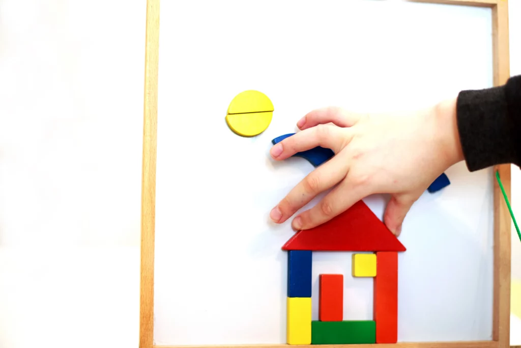 Child's hand placing colorful geometric wooden pieces on board forming a house and yellow circle.